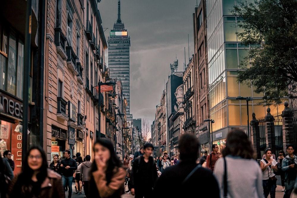 A still photograph of a busy Mexico City shopping street with countless pedestrians and high-rise buildings in the distance. Mexico City and its skyline is a popular backdrop for some top Mexican movies
