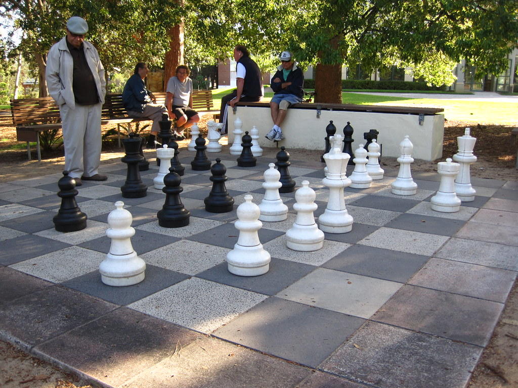 A life-sized chess board designed on the floor through tiles in the park. There are life-like chess pieces, and it looks as if an intense game is going on. Playing chess is a favorite pastime among older people, especially in countries like Russia and the United States