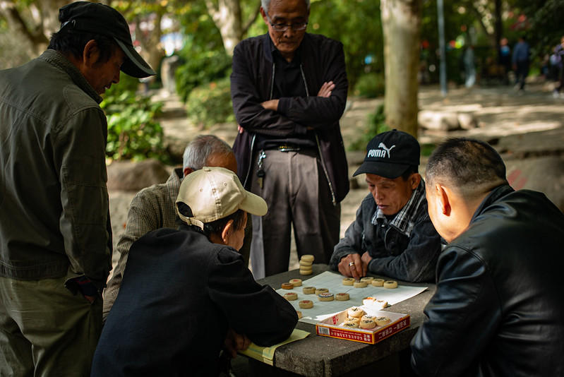 Chinese elderly playing Xiangqi, a variant of chess that became popular in China. Two people play the game while four are gathered around and watching the game being played on a stone table. If you have found yourself as part of a chess audience, you may be interested in taking chess lessons online