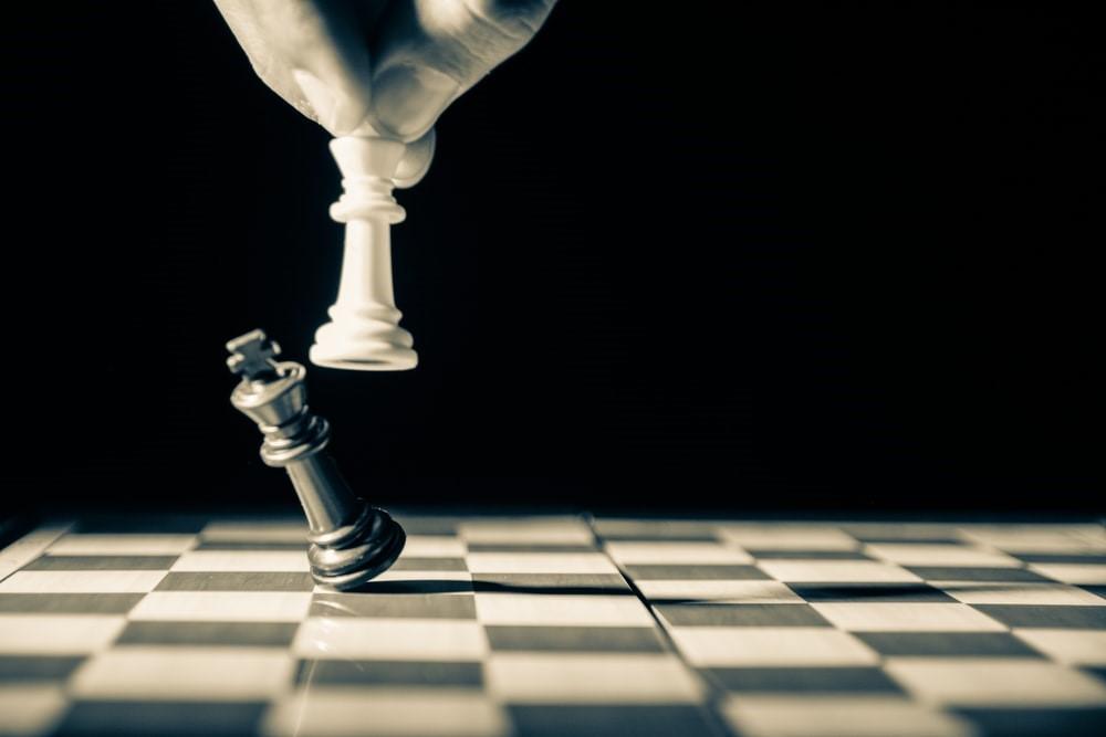 A white queen piece knocking a black king piece on an empty chessboard. Playing chess can be an exhilarating activity, especially during the end stages of the game, when you’re close to an inevitable checkmate