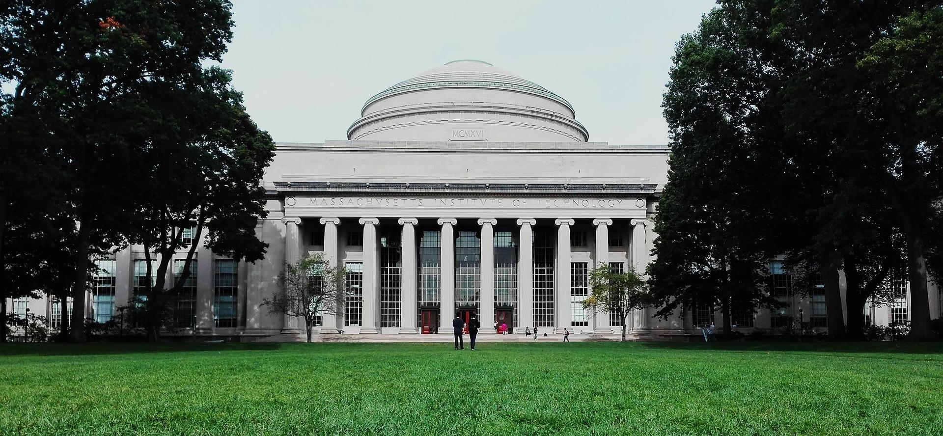 The Massachusetts Institute of Technology (MIT) front view shows two people walking towards the institute. Trees line both sides of the institute. 