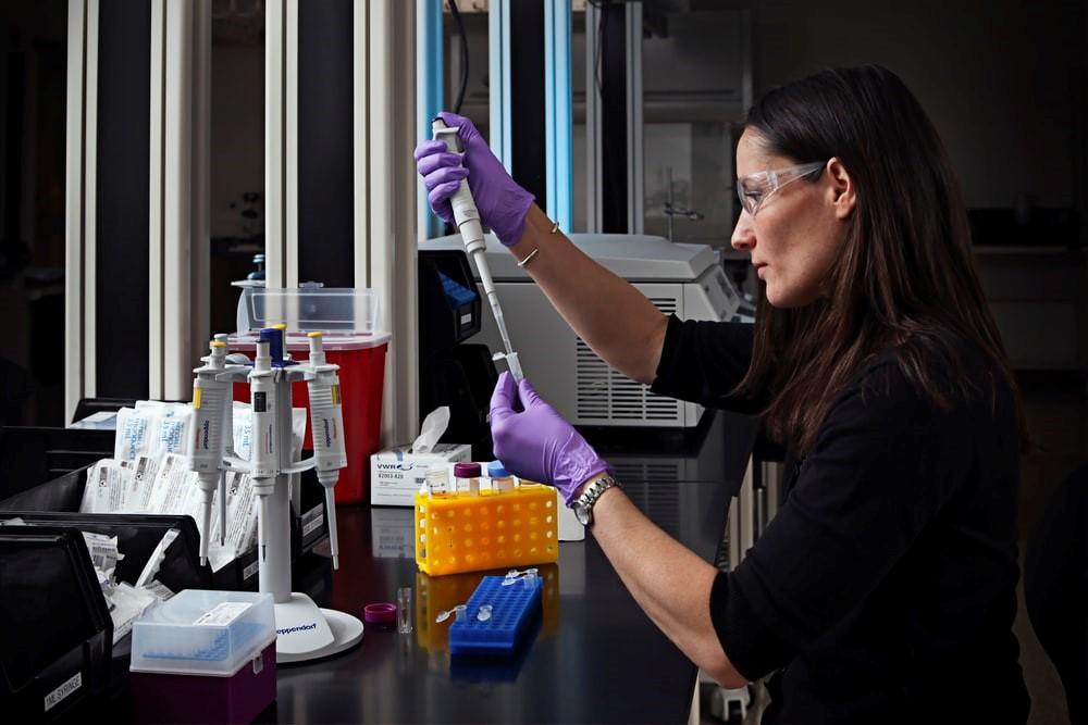 A girl wearing personal protective gear and using a micro-pipette to fill small centrifuge tubes. There is a stand holding multiple pipettes, a test tube and centrifuge tube stand, and a centrifuge in the background.