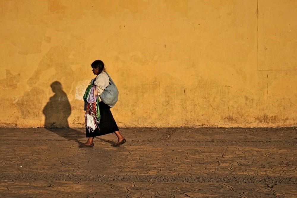 A woman in a white shirt and black skirt is walking past a yellow wall. Mexico is renowned for its unique architectural features, such as adobe walls. Studying Spanish in Mexico will help you gain an authentic knowledge of the architecture of the country.