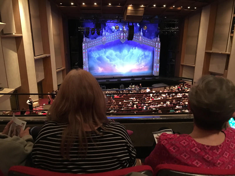 Audience members are seated in a Broadway theatre putting on The Book of Mormon musical. Everyone has been handed their playbills before the show starts to know what will happen in the program