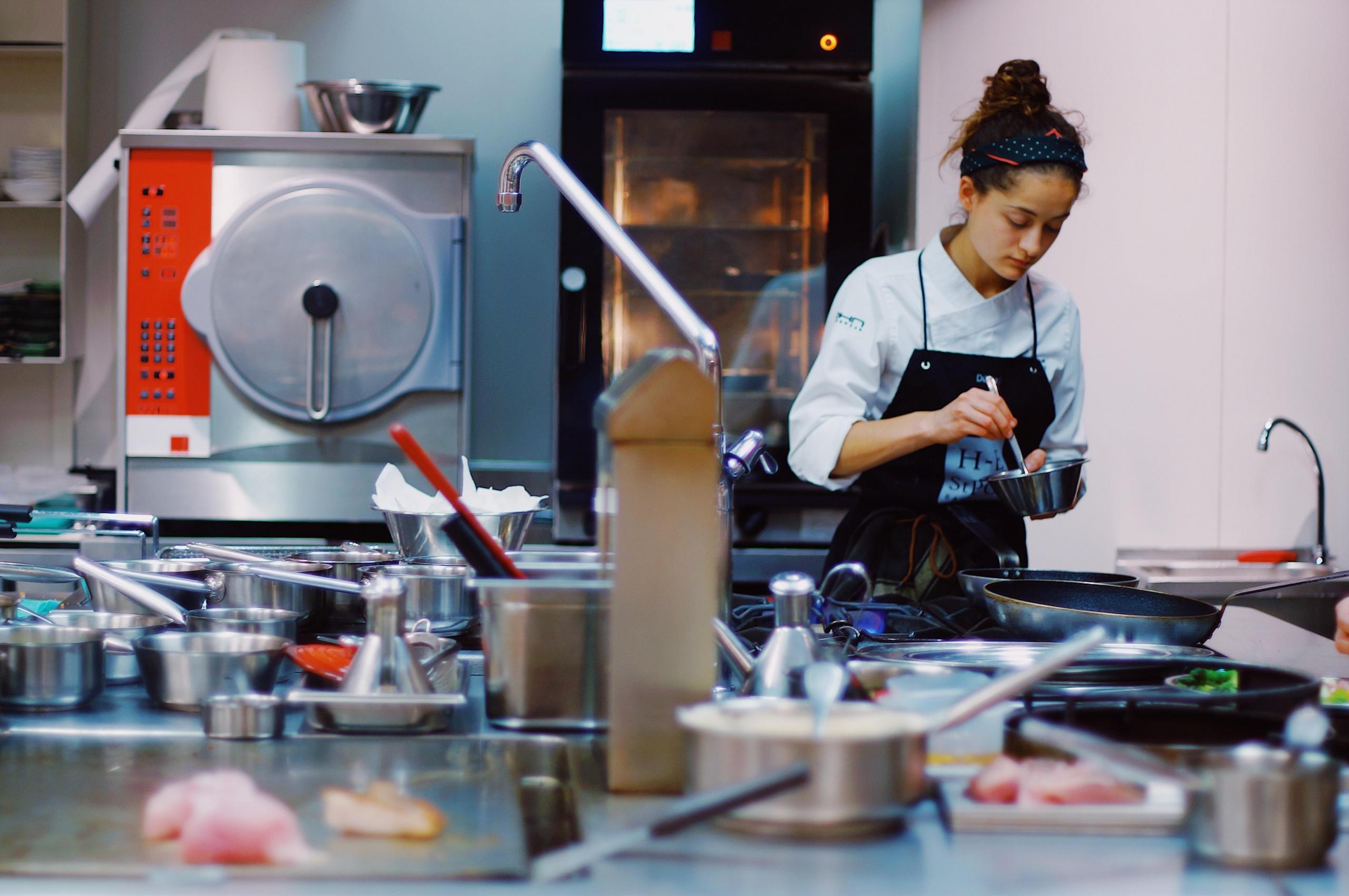 A female chef holding a utensil and mixing something; several steel utensils are also in the room. All the top chefs in the USA started from scratch, taking it one day at a time towards their ultimate success
