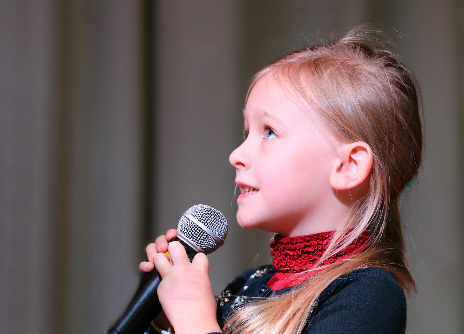 A little girl is holding a microphone and singing. She is wearing a black and red dress with tiny white pearls. Training your voice before a performance can prove to be fruitful if you have to perform in front of many people on stage