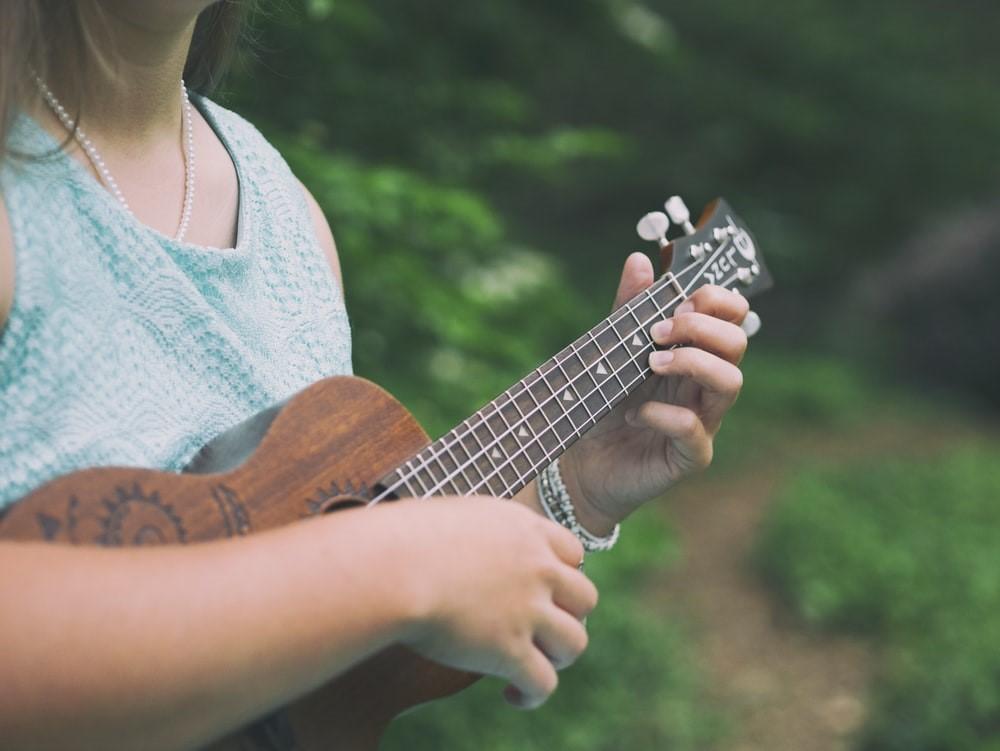 A woman in a green shirt wearing a pearl necklace and holding a guitar in her hand as she strums it. It is hard to train your voice without the help of a voice coach or a musical instrument to act as a guiding base.