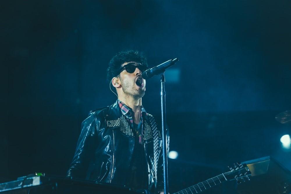 A band vocalist wearing sunglasses and a leather jacket holding a guitar and singing into a microphone in a dark place. Most of the famous musicians take regular voice training classes to maintain their singing quality and tune