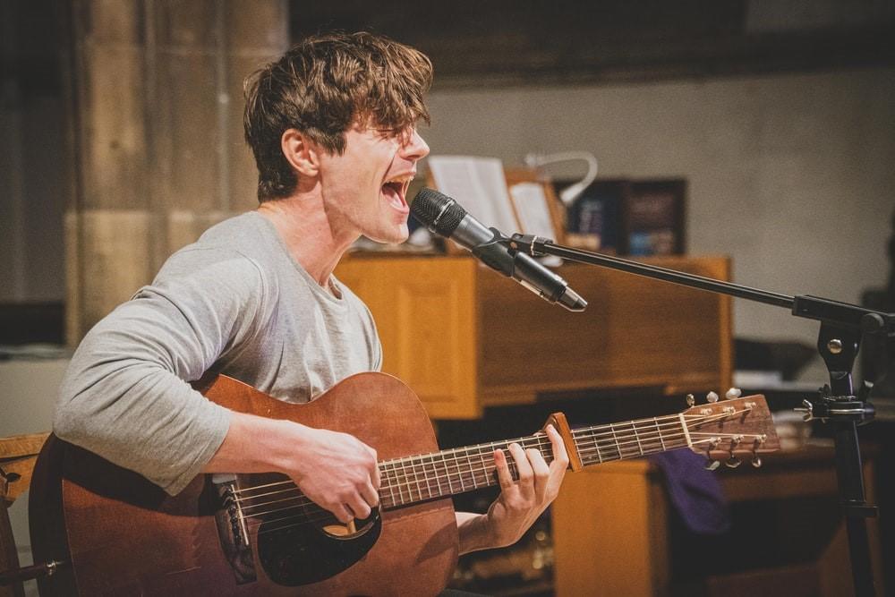 A man in a grey shirt holding a guitar and singing into a mic with a wooden piano placed behind him. Singing in front of a live audience can be straining, but even singing for pre-recorded sessions can be stressful. Get past these problems by training your voice!