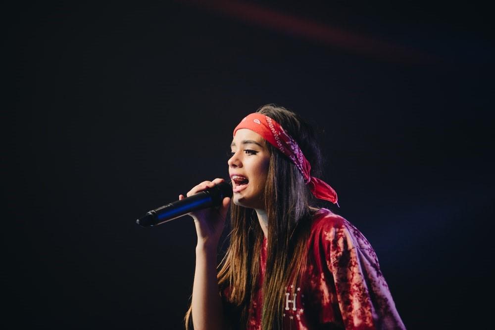 A woman in a red shirt and a red bandana singing into a microphone she is holding. An essential part of voice training involves projecting your voice over a considerable distance; however, that is not the same as screaming into a mic