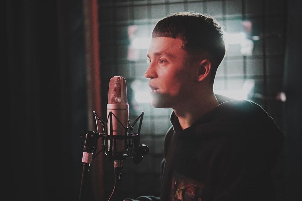 A man in a grey hooded sweatshirt singing in a mic in a dark room with a light in the background. Voice training is something all singers need to go through if they aspire towards a professional career
