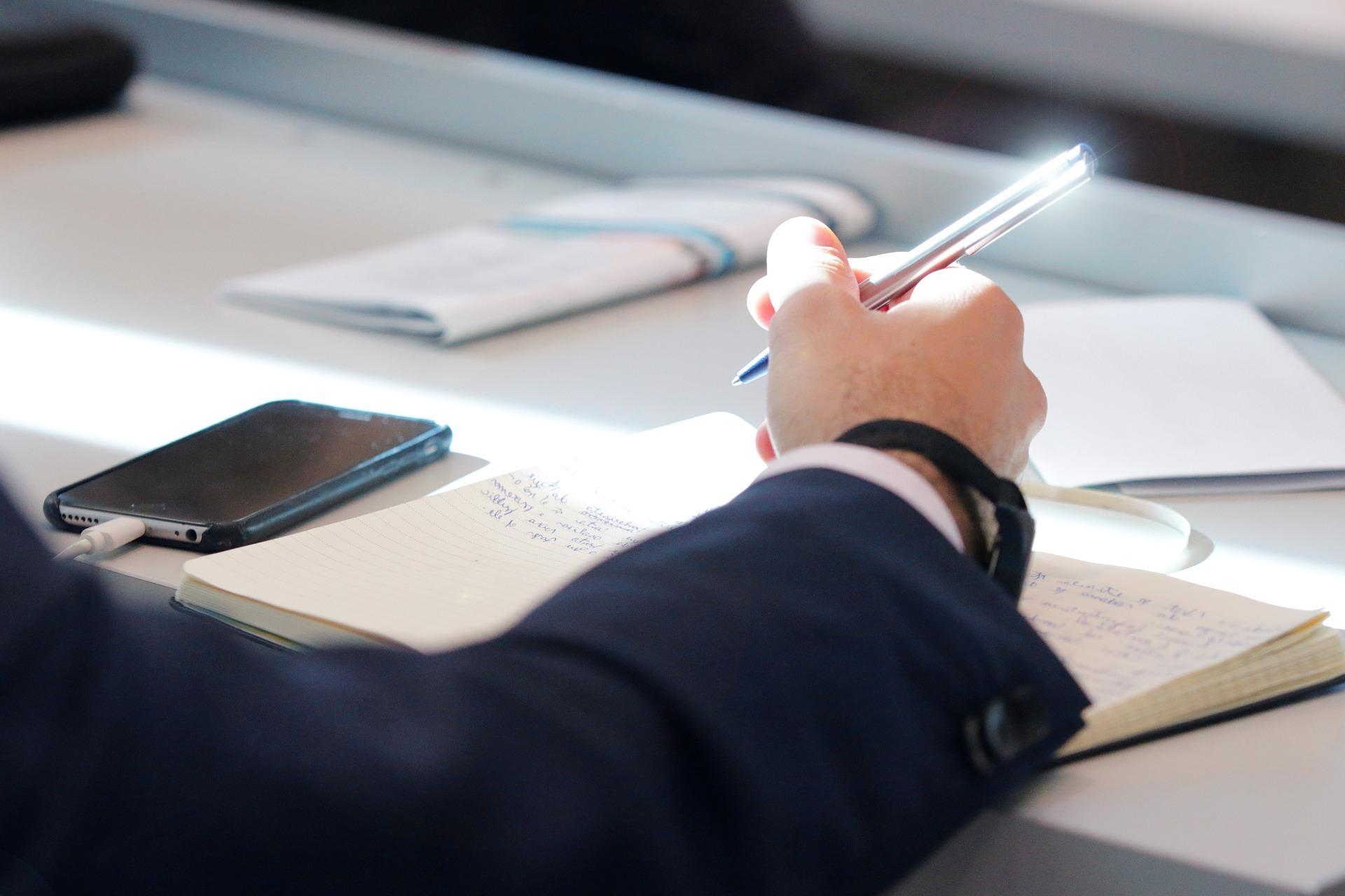 A guy wearing a blue coat and writing something on a notebook with a blue ballpoint pen. His phone is also plugged into a charging port beside him. Taking an AP French exam requires a lot of practice