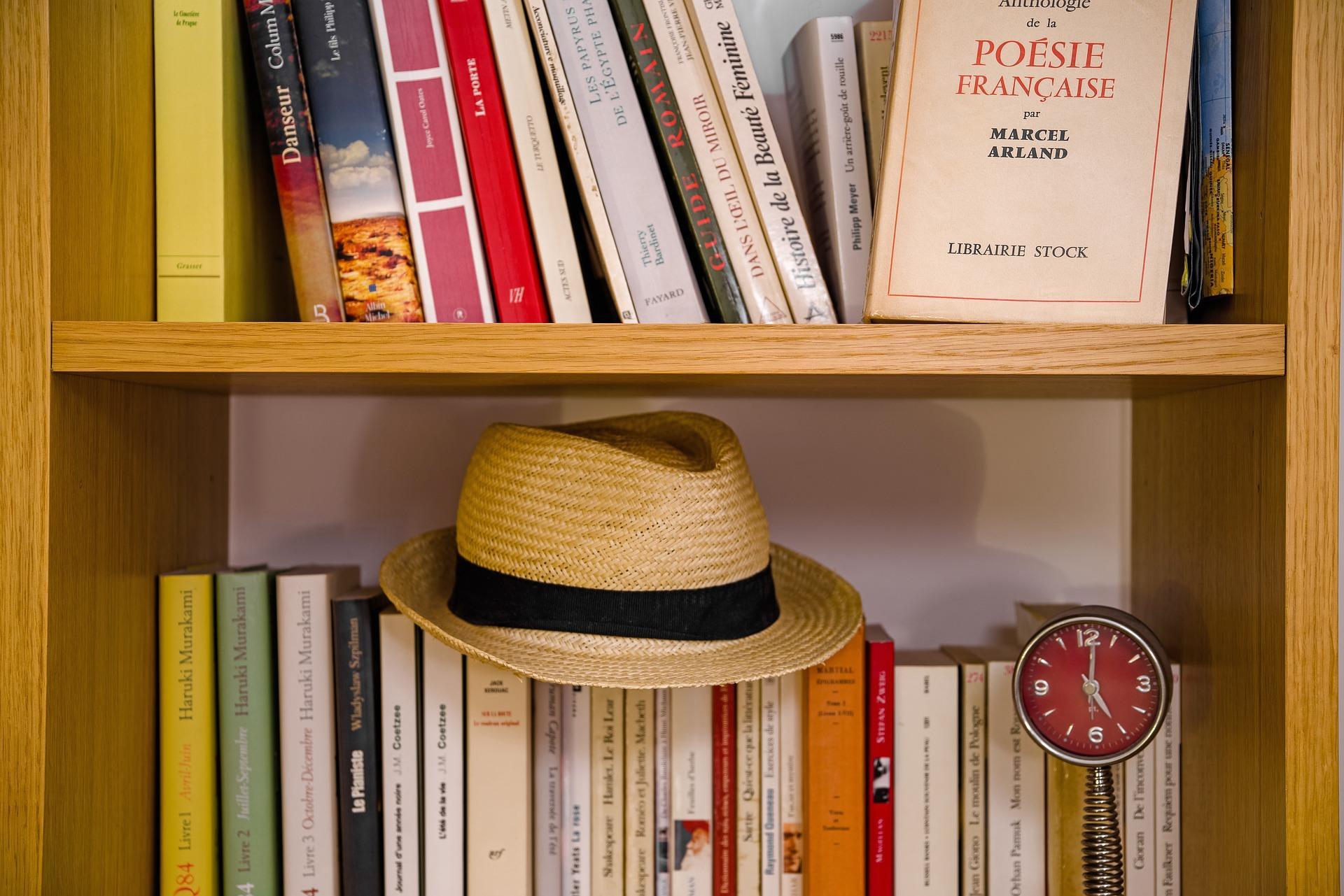 French books lined up on a wooden shelf. The most prominent book on the top shelf is of French poems. On the lower shelf, a straw hat and an ornamental clock are casually kept. The AP French test focuses on testing communication rather than grammar
