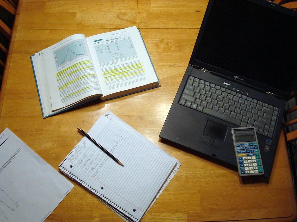 A laptop, calculator, math notebook, notes, and pencil spread out on a wooden table