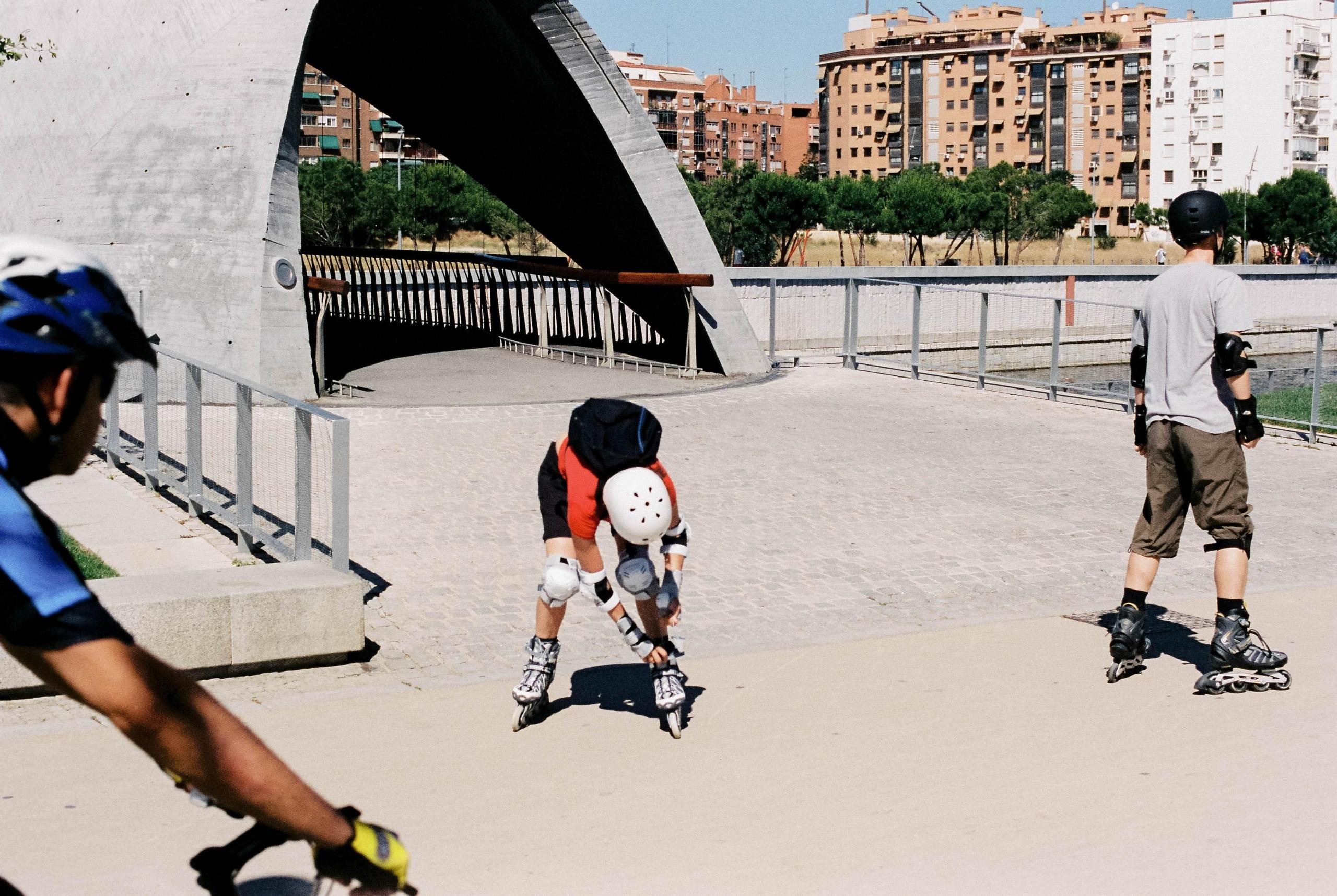 A guy in a gray shirt rollerblading while another one in an orange shirt and black bag is tightening his rollerblades