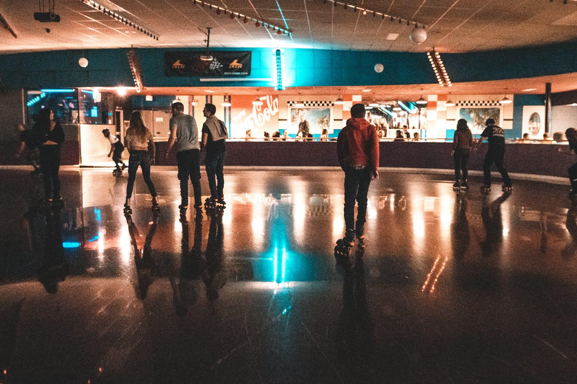 People roller skate across an indoor rink floor under bright blue and orange lights.