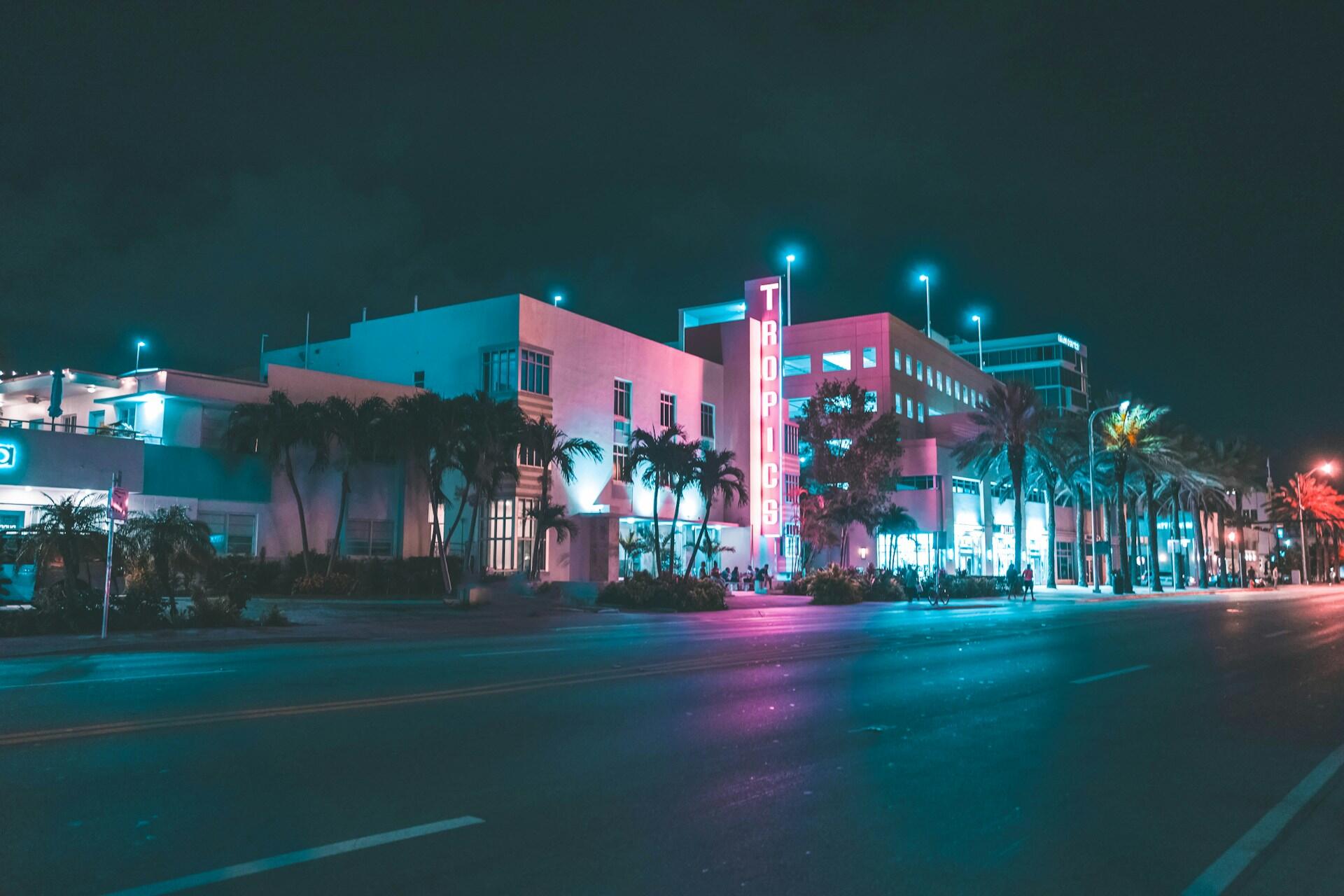 Neon-lit street scene with a building sign reading Tropics and palm trees at night.