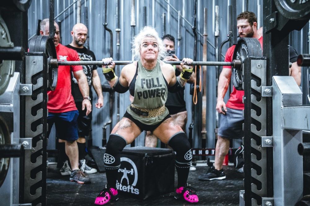 A woman lifts a barbell at a gym while her trainers keenly watch her do it