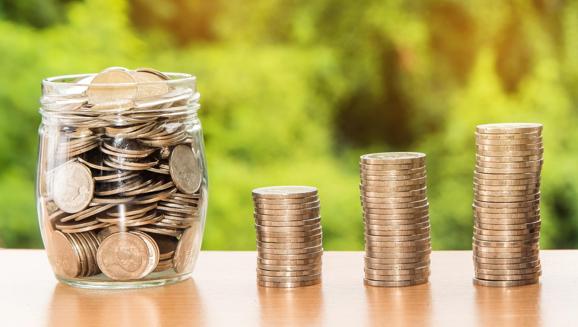 Coins neatly stacked up on a table and a jar full of coins next to the three stacks