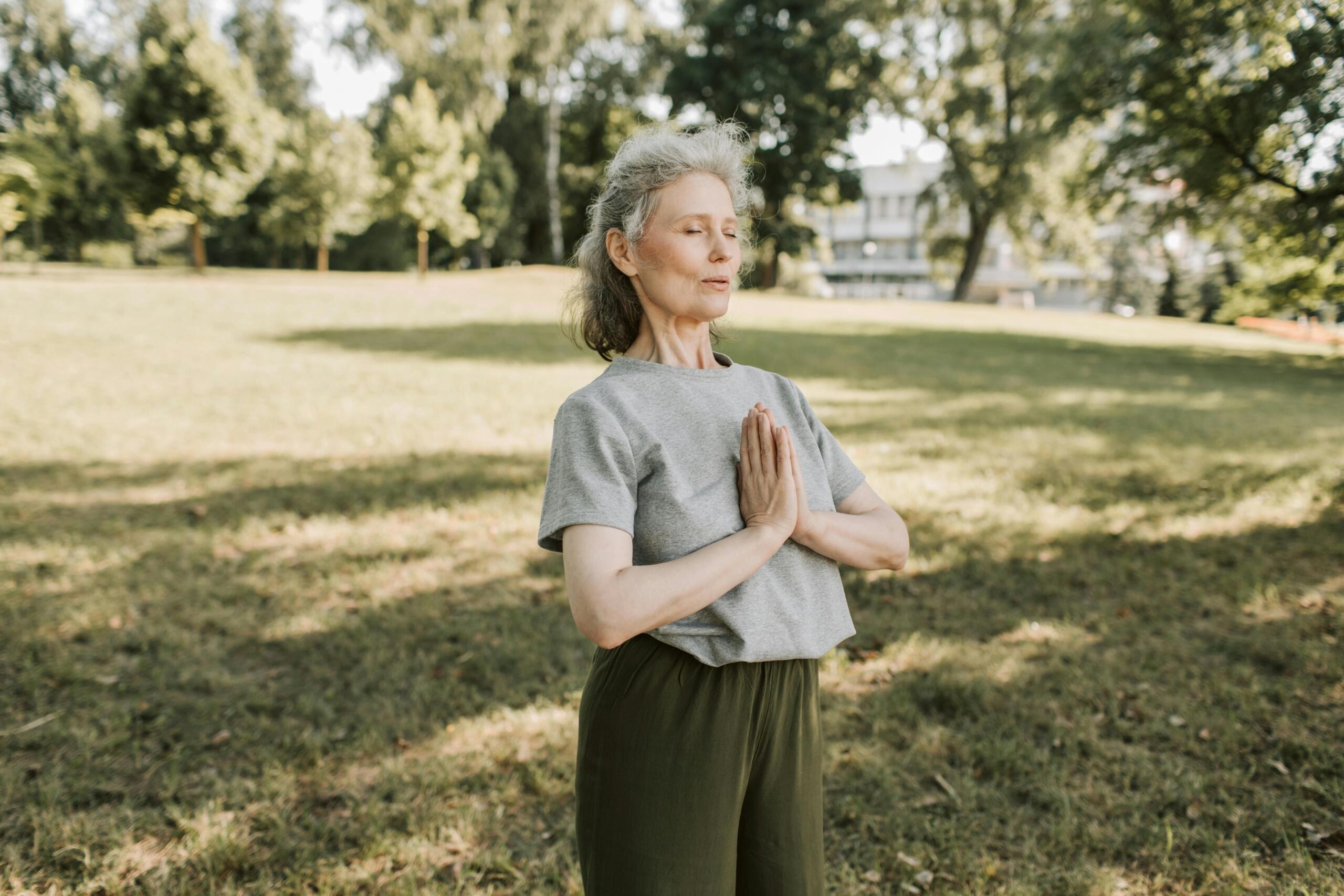 An elderly woman in a gray shirt is meditating peacefully at the park, surrounded by greenery and natural light.