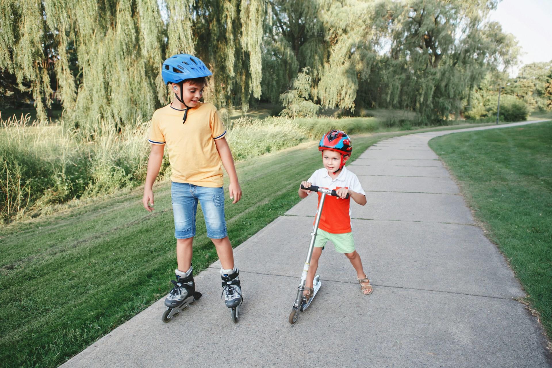 Two kids on a trail outside learning to roller skate and ride a scooter.