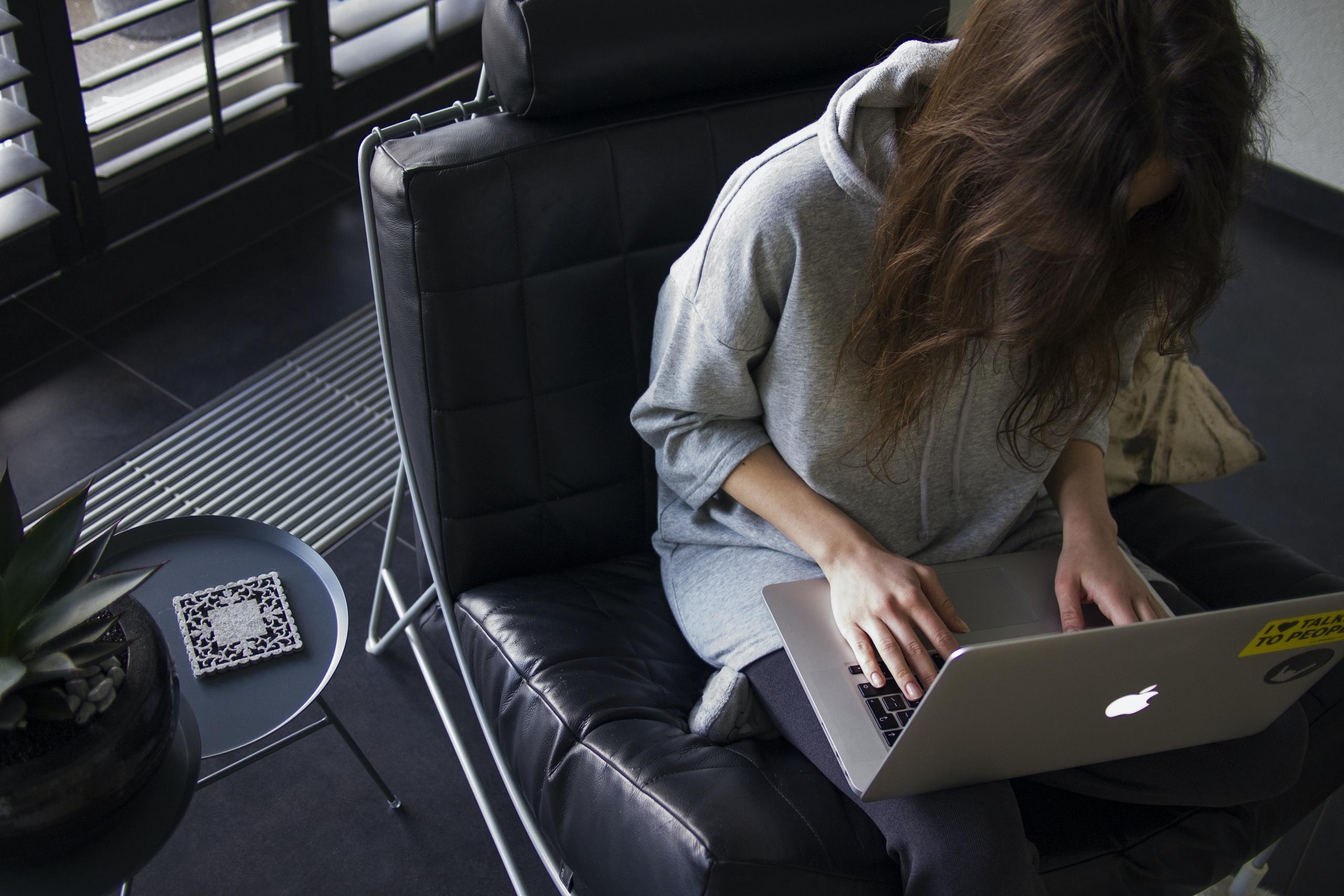 A woman in a light-blue chambray long-sleeved top sitting on a black leather sofa while writing her personal statement on the laptop