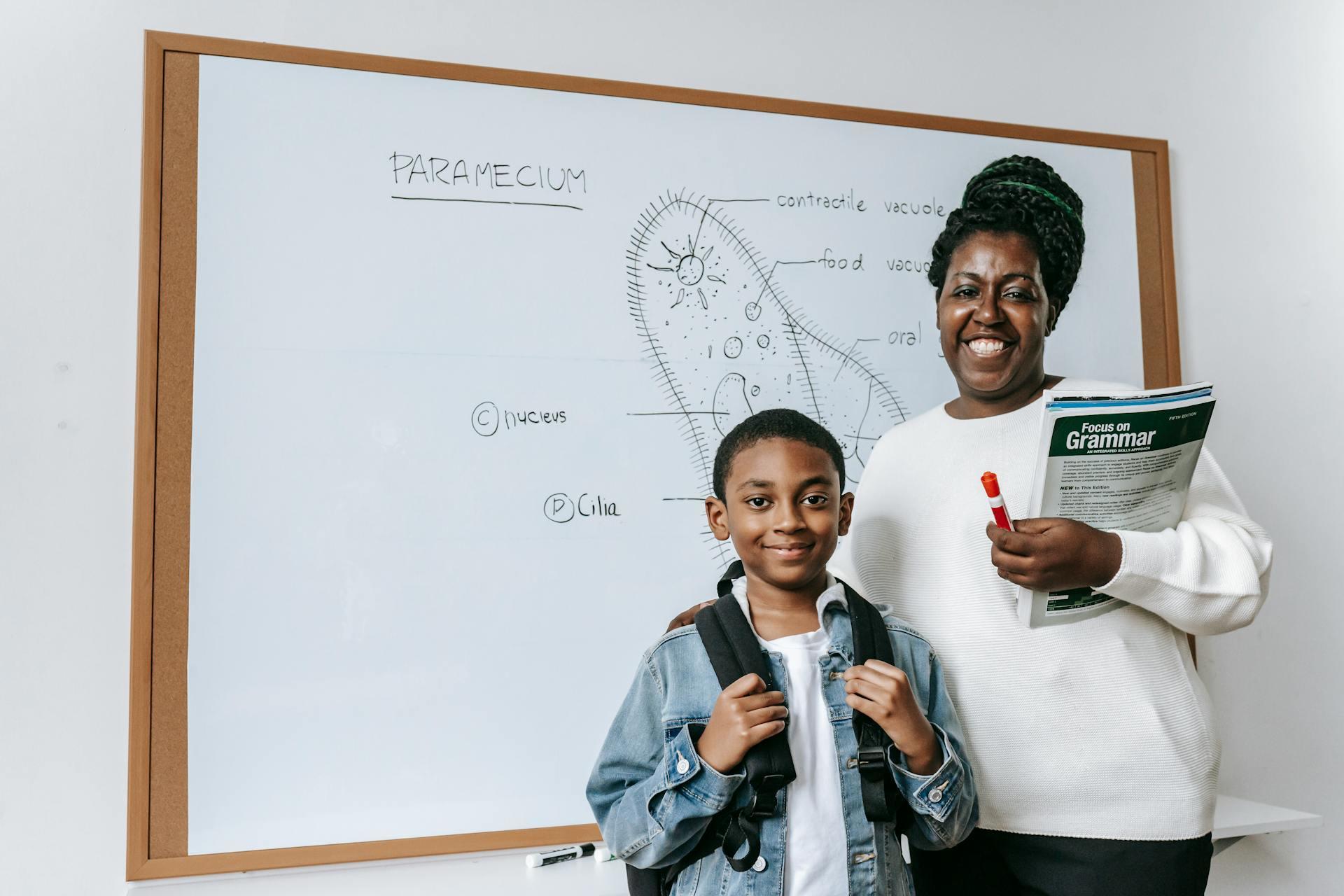 A teacher and young student standing together smiling in front of a whiteboard with a diagram of a paramecium drawn on it