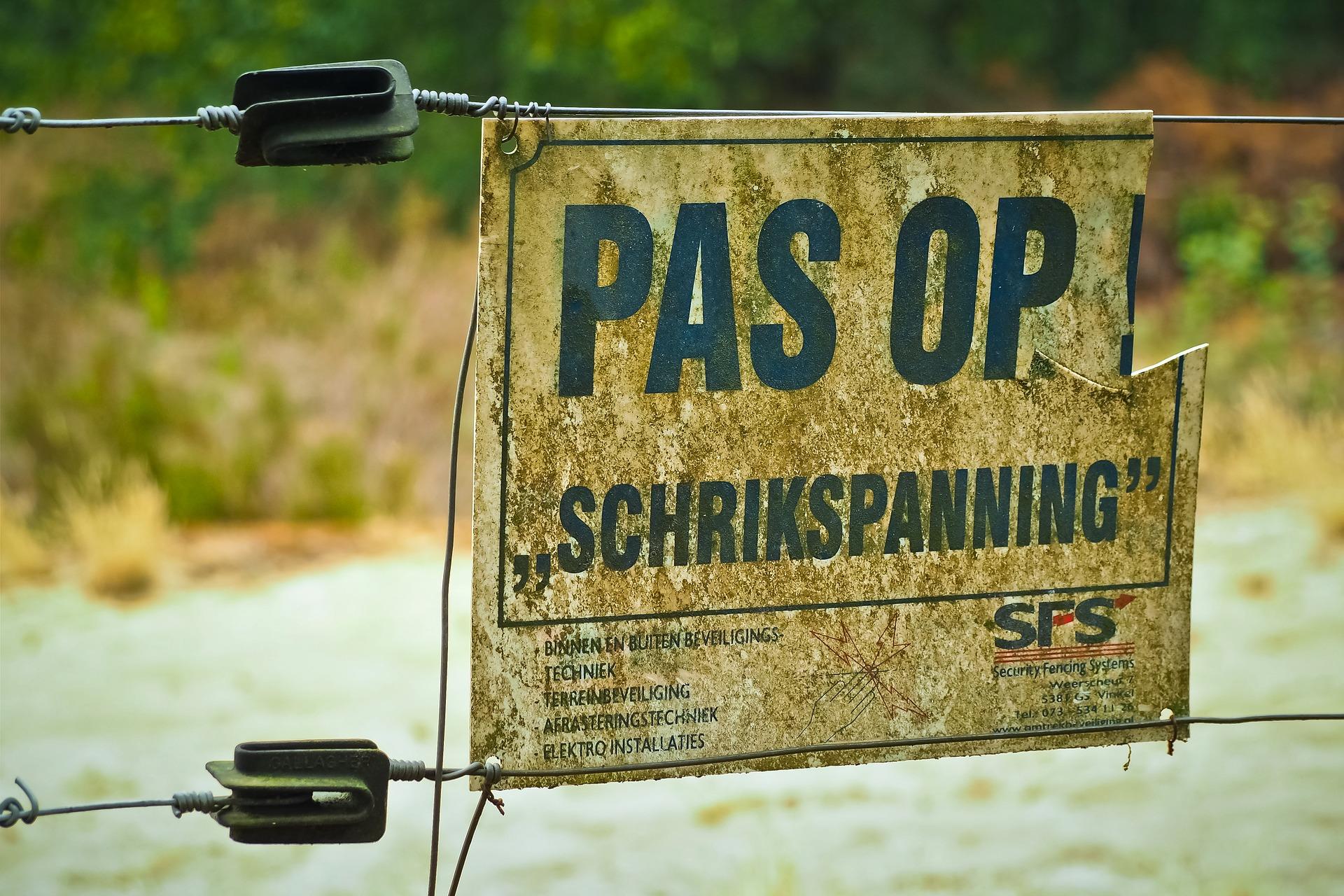 A warning sign outside the military field saying "Pas Op Schrikspanning," which means beware of electric shock.