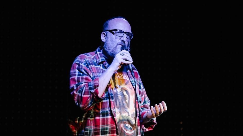 A man in red, white, and blue plaid dress shirt performing standup in NYC