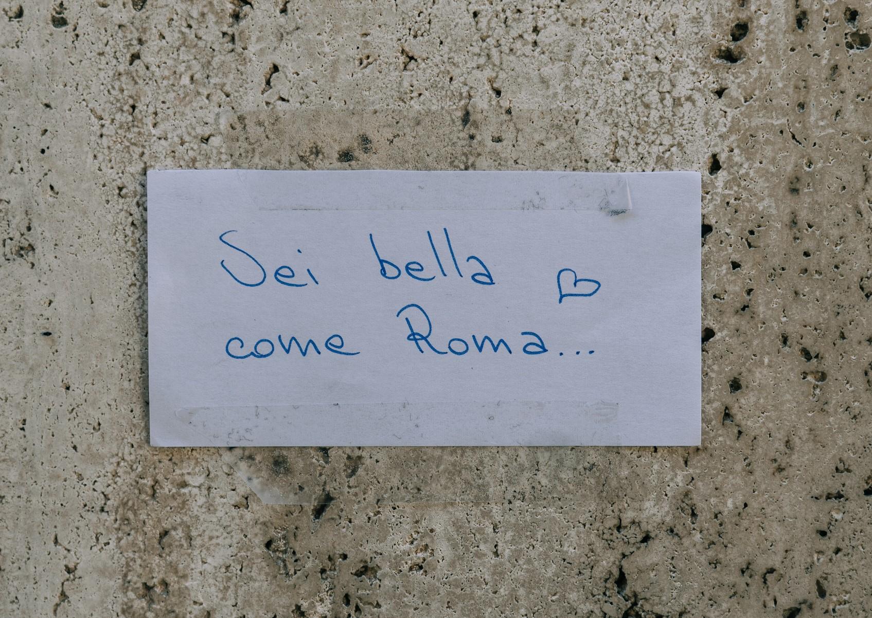 An Italian handwritten note taped to a wall, saying "You're as beautiful as Rome" in a Roman street