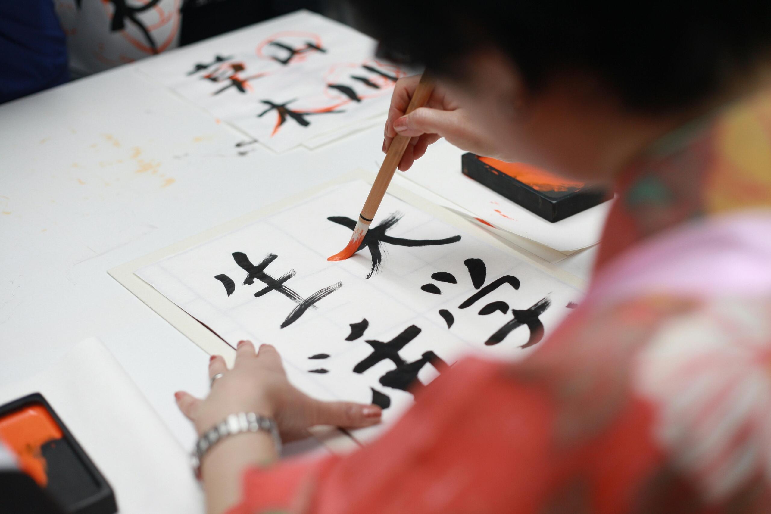 A person holding a brush while drawing Kanji script on paper.