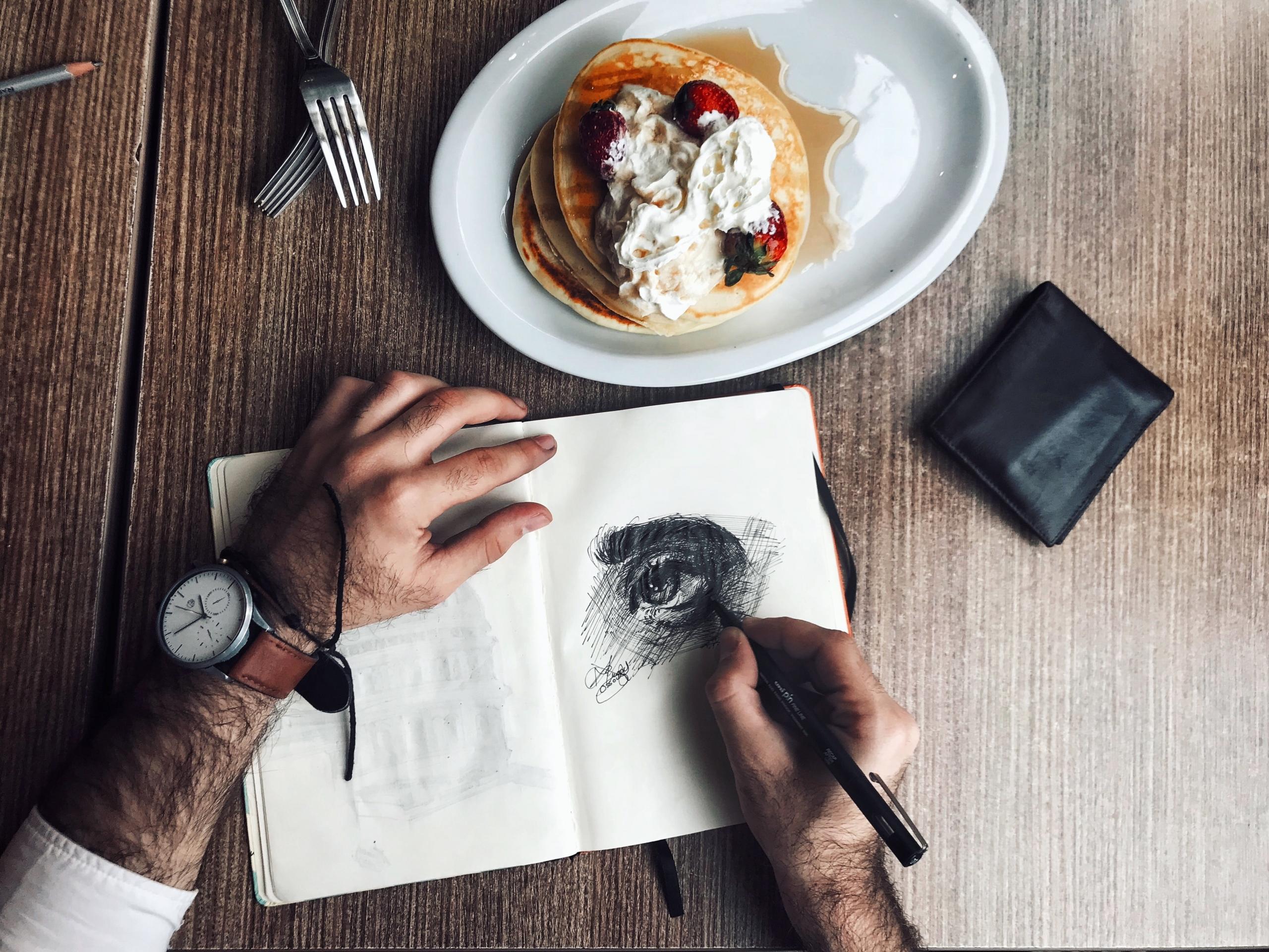 A man sketching an eye in a notepad, with a plate of pancakes next to it