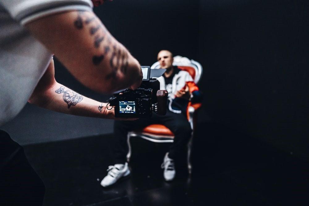An actor sitting on a chair as the tattooed cameraman records him