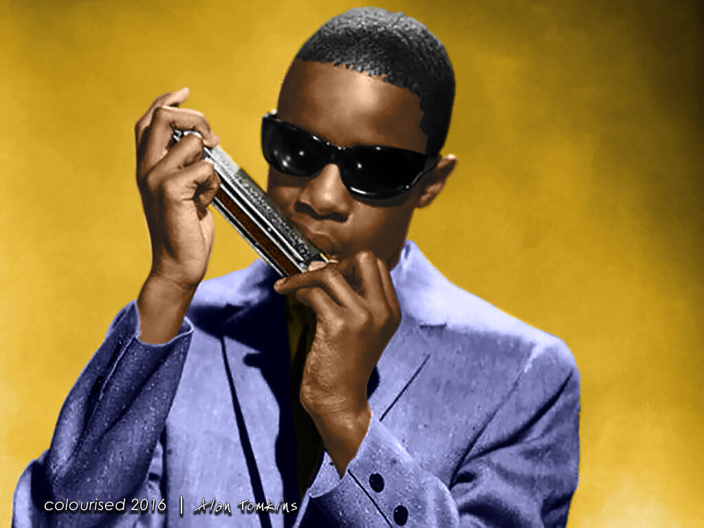 Stevie Wonder wearing sunglasses and a blue suit while playing the harmonica.