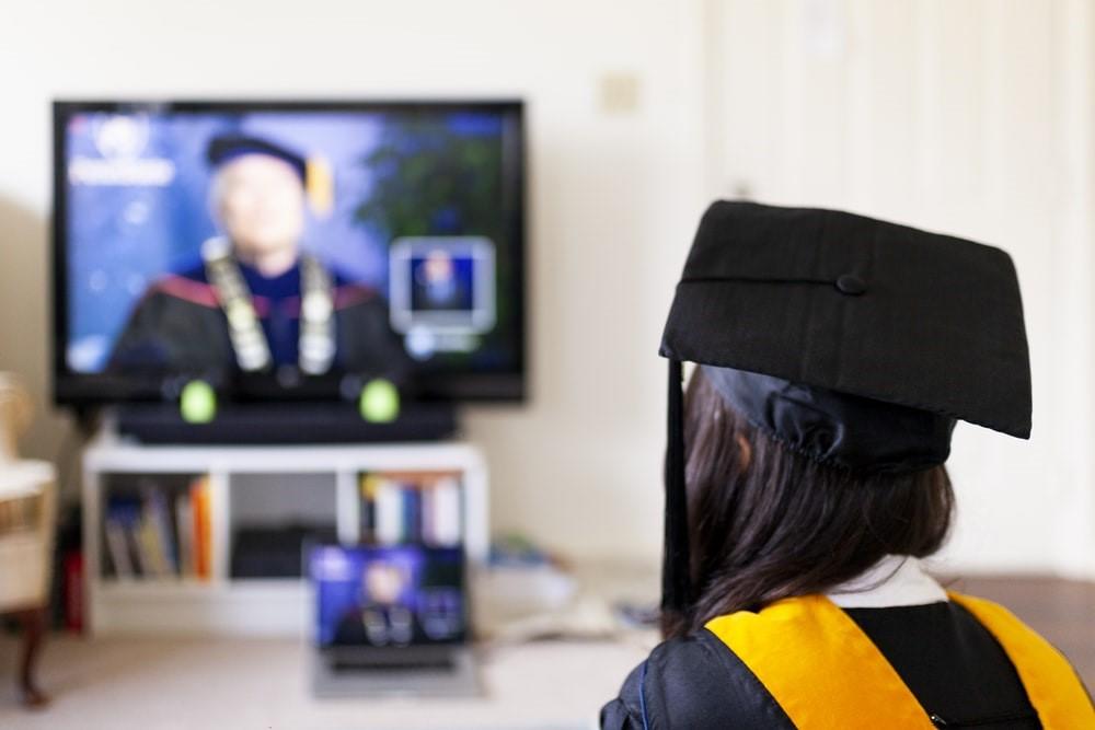A freshly graduated student watching a live commencement ceremony on her TV