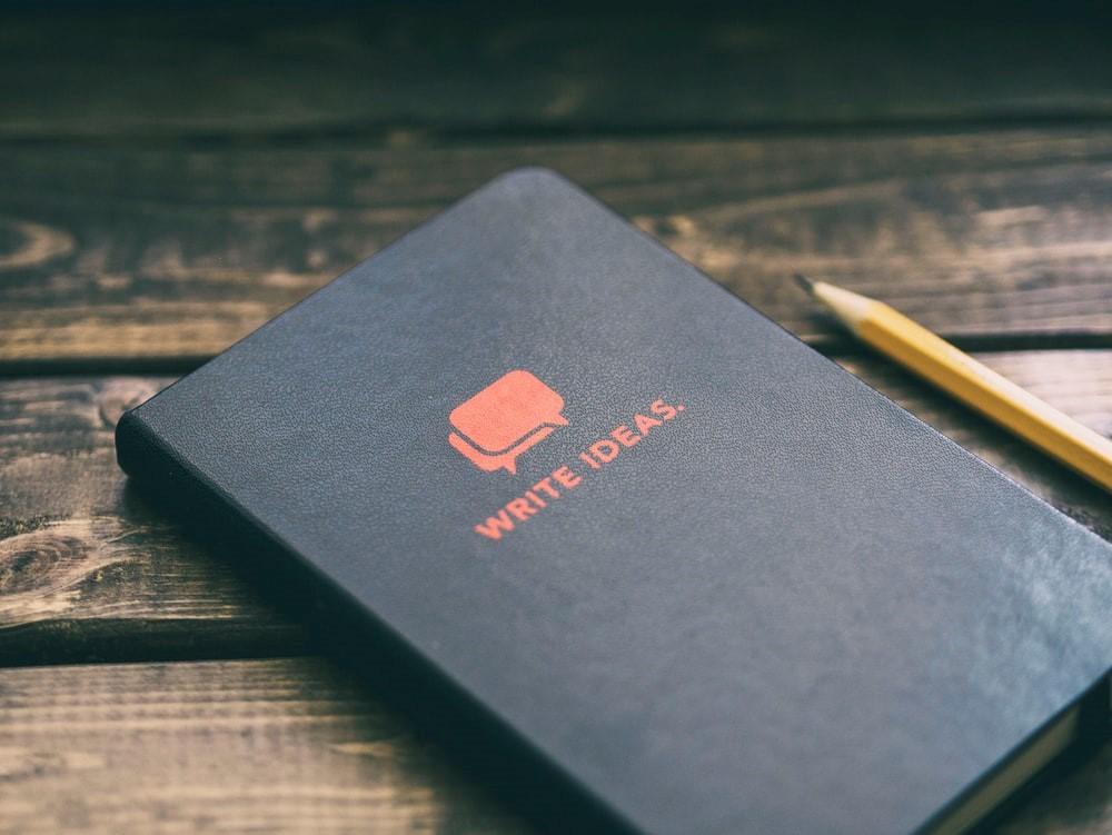 A notebook placed on a wooden table next to a pencil with 'WRITE IDEAS' written on the cover