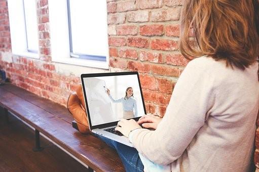 A blonde woman is taking online classes on her laptop while sitting on a bench next to a window