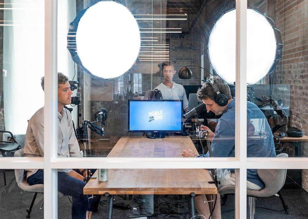 A journalist interviewing a person in a studio for a podcast with a monitor on a brown wooden table and studio lights surrounding it.