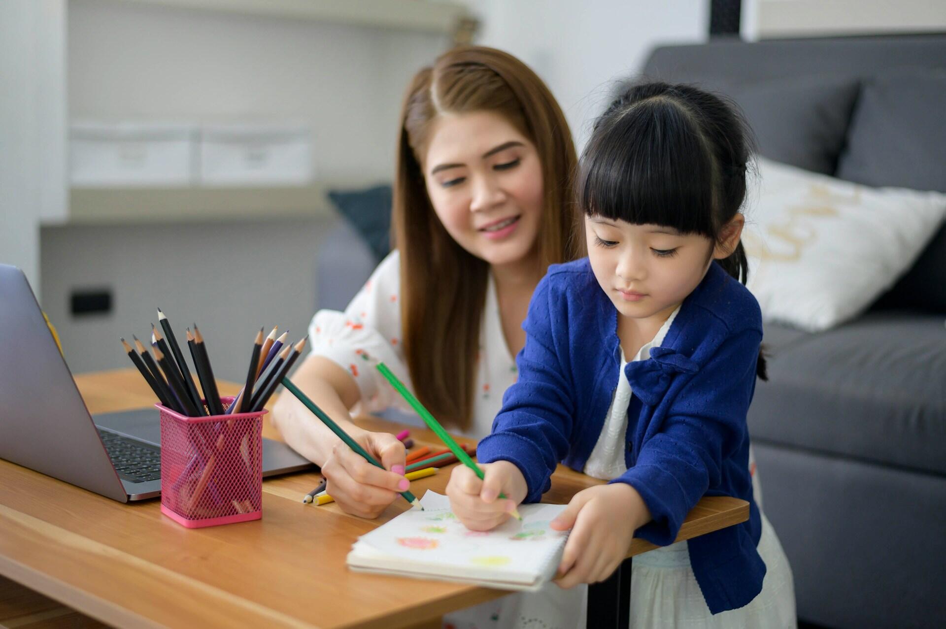 A little girl coloring on a notepad with a tutor behind her helping.