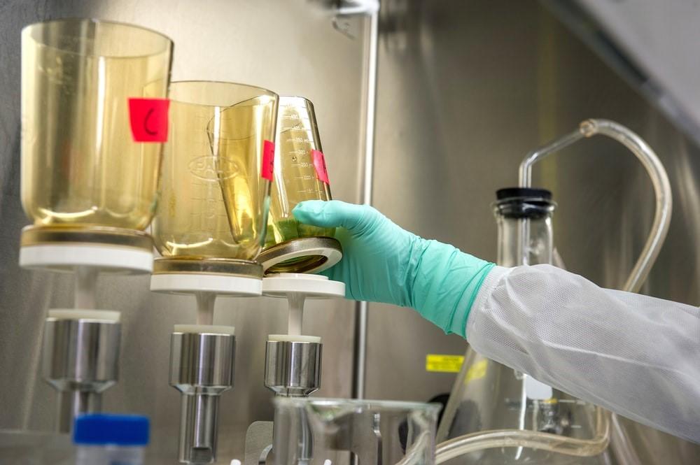 A person with a green disposable glove is holding a transparent golden flask with the letter "A" on it and various other flasks around in a lab
