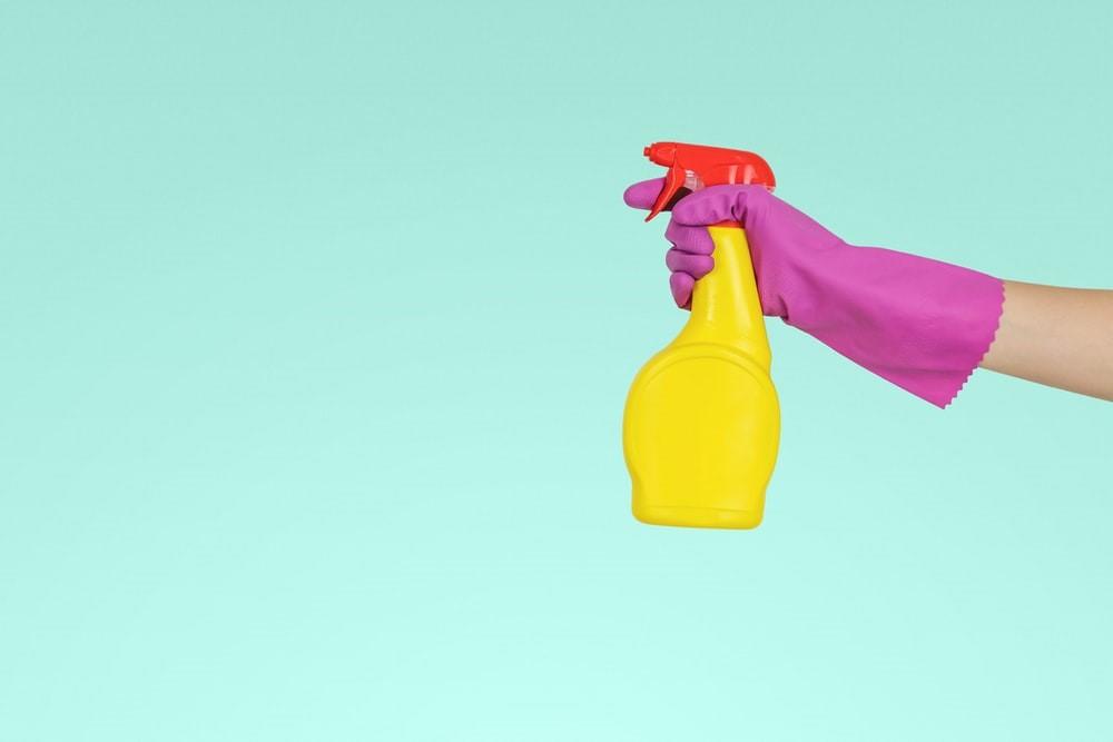 A person holding a yellow detergent spray with a pink glove on against a turquoise background
