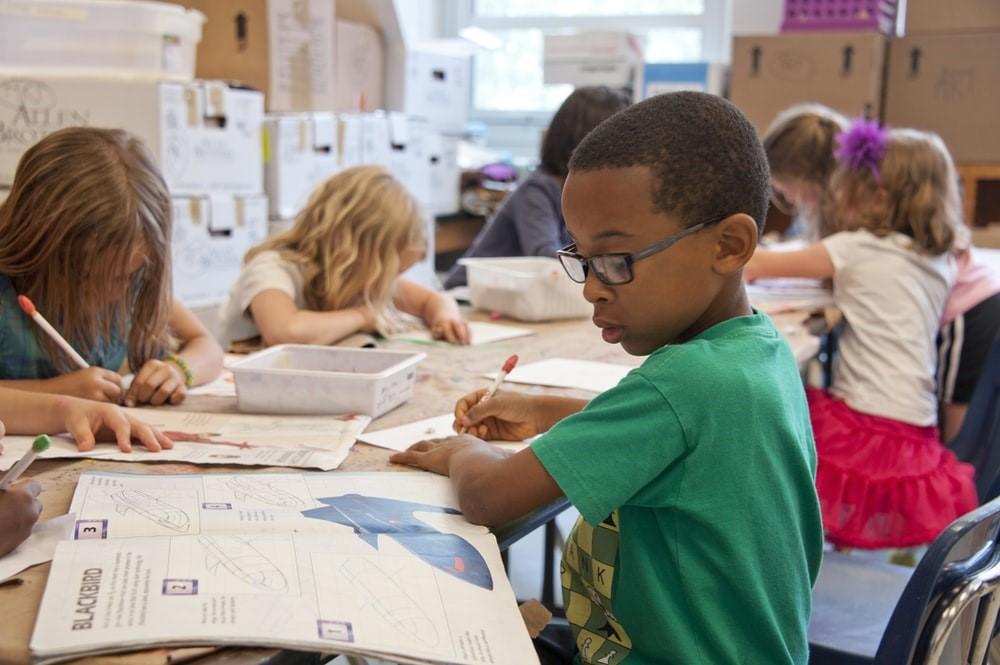Kids wearing casual clothes with pencils in their hands sitting on a long desk next to each other with banks on the desk.
