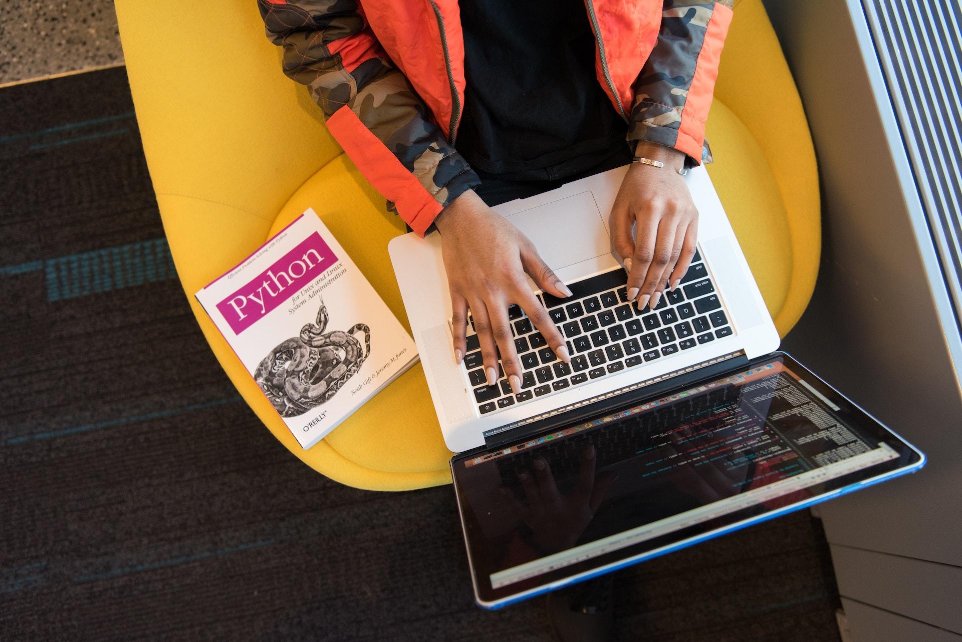 A person sitting in a bright yellow chair has a laptop on their knees and a Python programming textbook close by