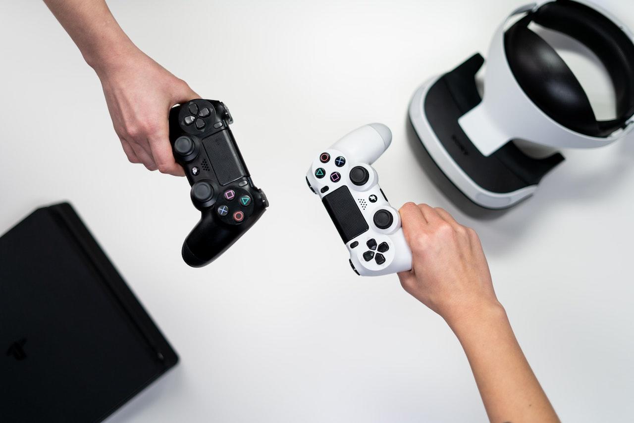 Two people hold Playstation controllers with a virtual reality headset in the background