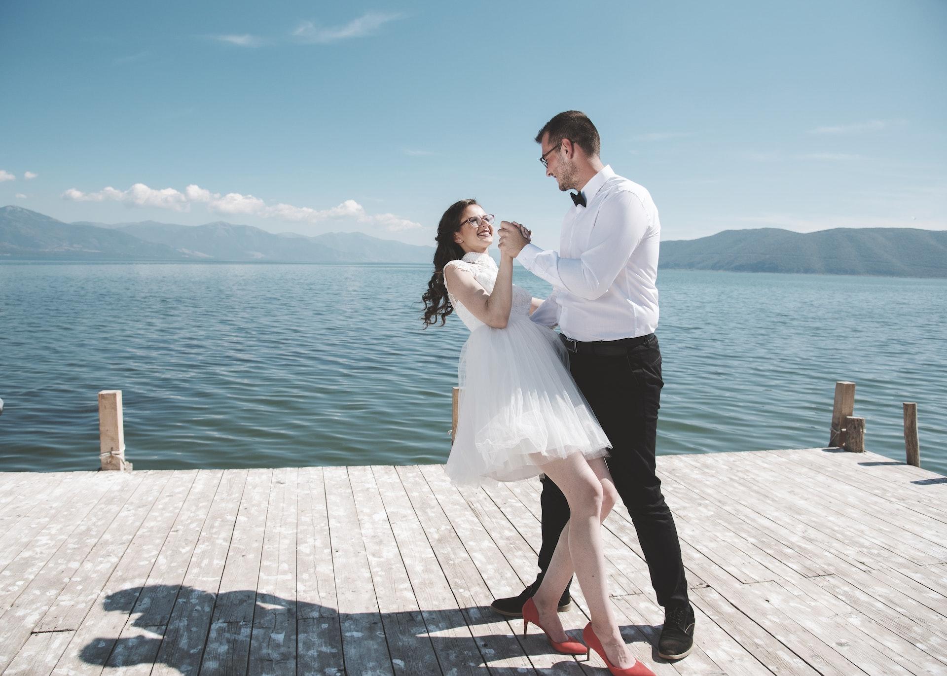 A newlywed couple in their wedding attire dancing on a dock near a lake.