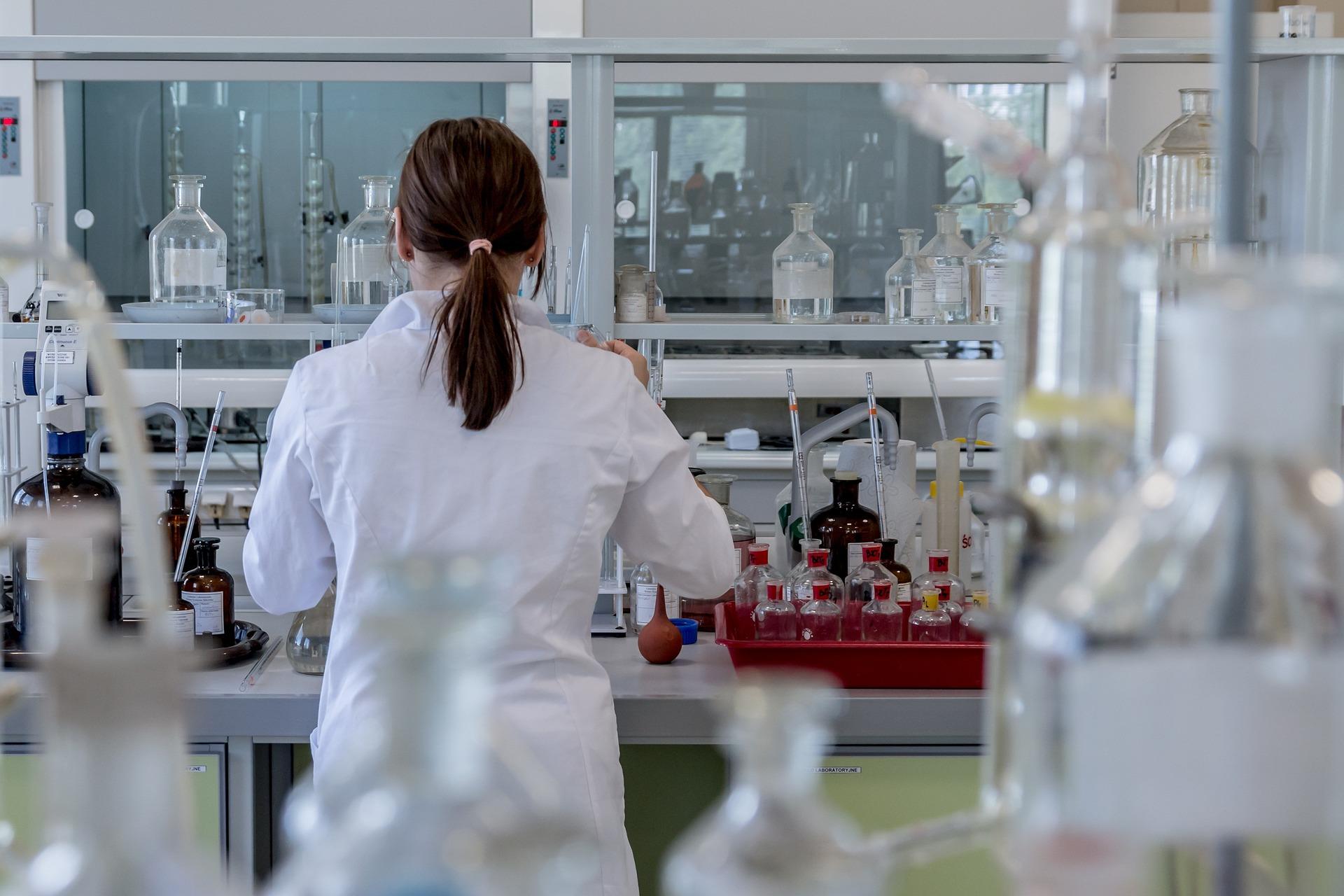 A scientist standing at a lab bench with her back turned to the camera, surrounded by glass testing vessels.