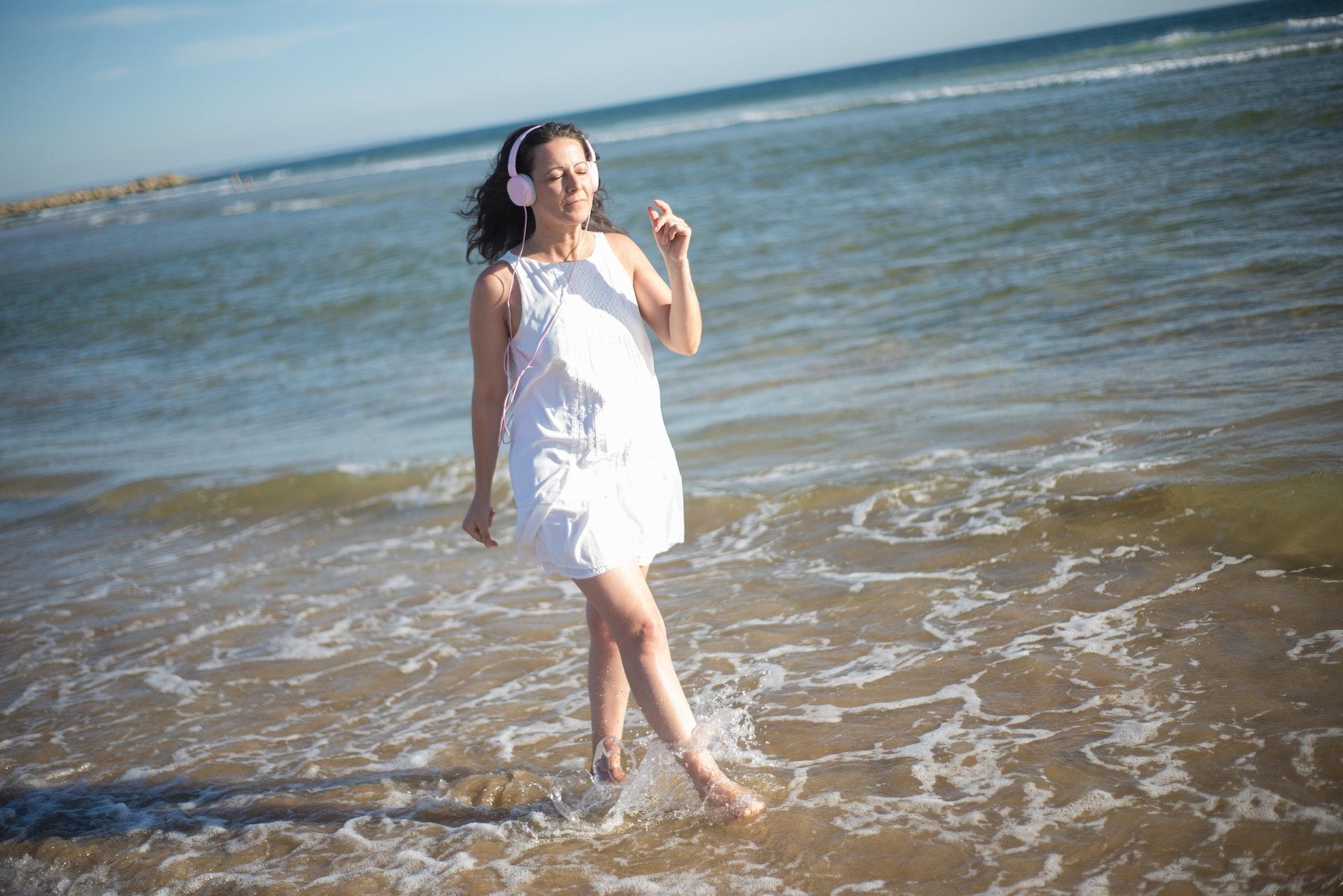 A woman walking in the surf at the beach and listening to music with headphones.