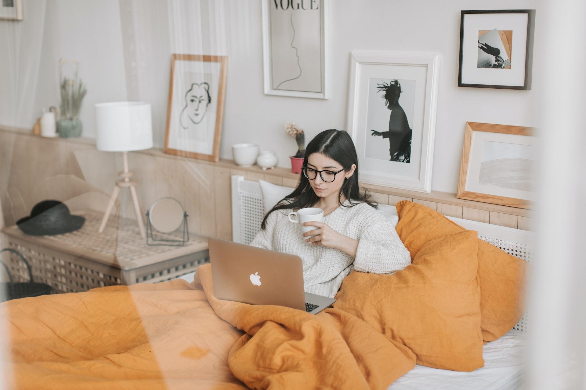 A woman watches a movie on her laptop while sipping tea and snuggled in her blankets.