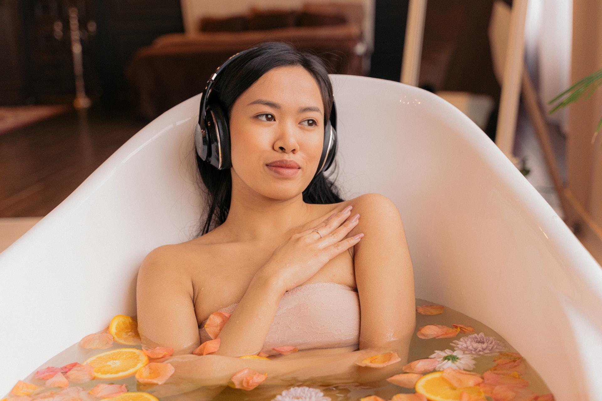 A woman sits in a bathtub with headphones on to listen to a podcast.