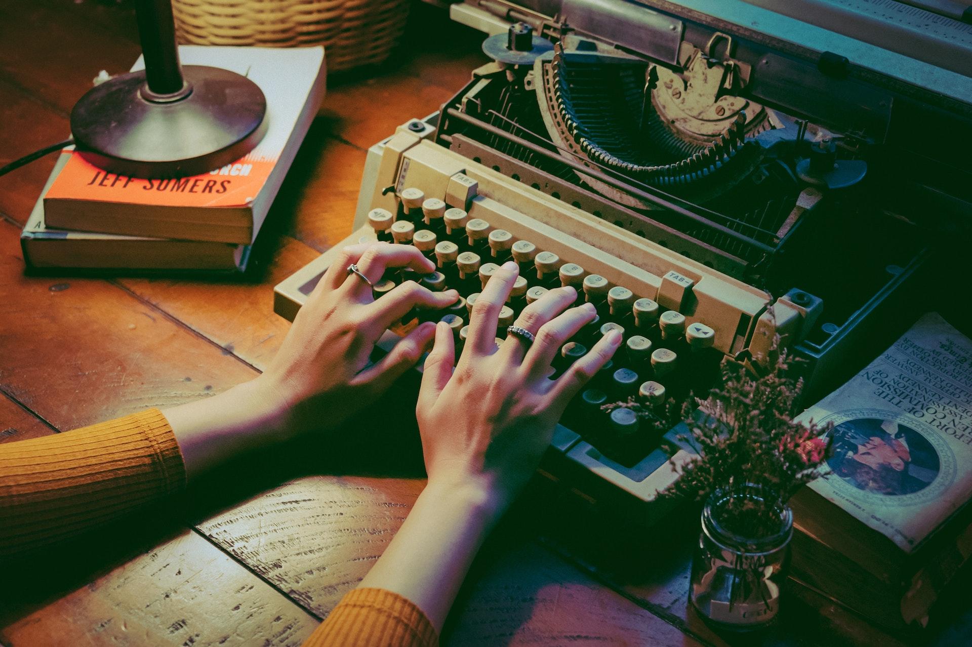 A pair of hands types on a typewriter that sits on a desk with flowers and books nearby.